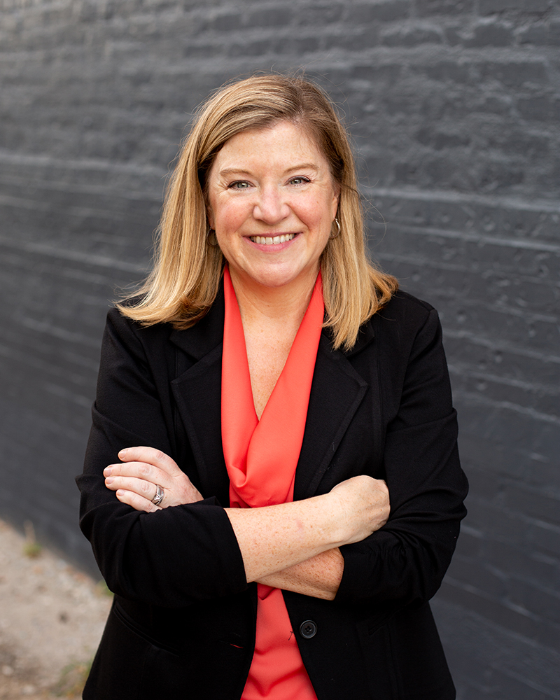 Jeannie Walters in front of a black brick wall, smiling with her arms crossed.