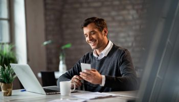 happy businessman texting on mobile phone while working on laptop in the office